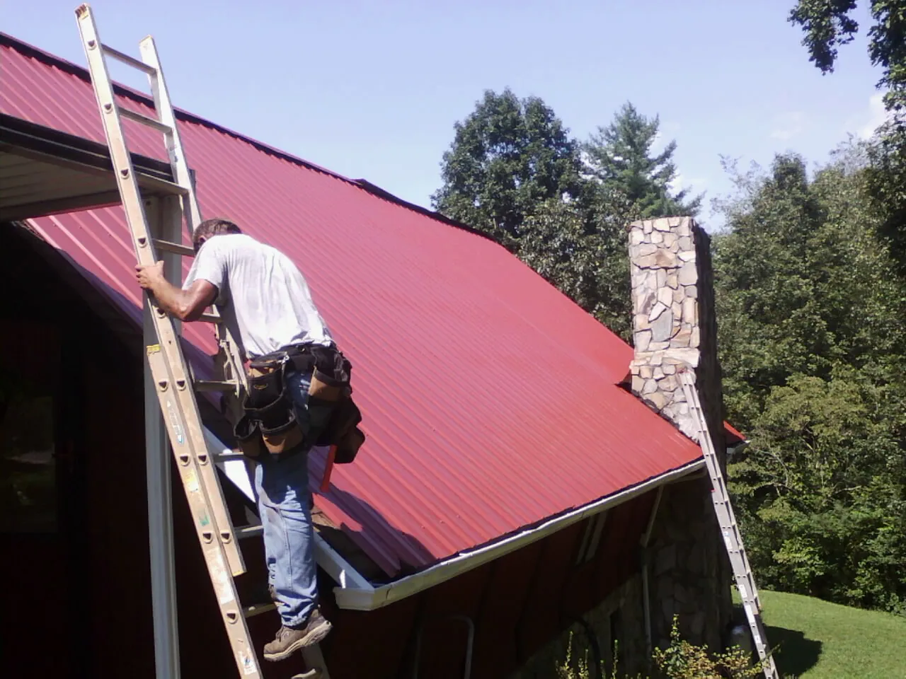 Roofer on a ladder beside a red metal roof and stone chimney — Soffit & Fascia Repair by Roofing Brothers in Cecil